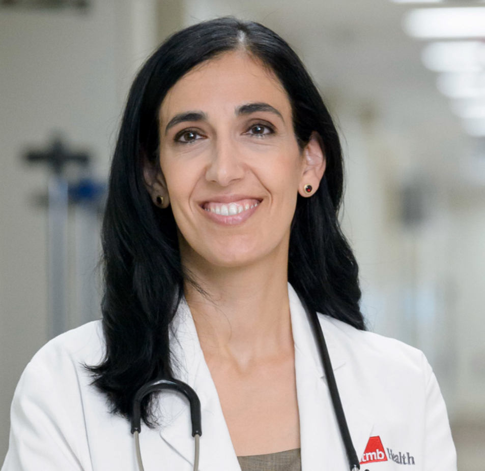 image of woman with dark, shoulder length hair smiling. she's wearing stud earrings, a white coat and a stethoscope around her neck. the image only displays until her chest. 