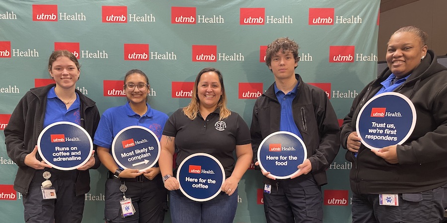 4 women, 1 man standing in front of a UTMB Health backdrop holding round signs that say "UTMB Health" and things like "runs on coffee and adrenaline" "most likely to complain" "Here for the coffee" "Here for the food" "Trust us, we're first responders"