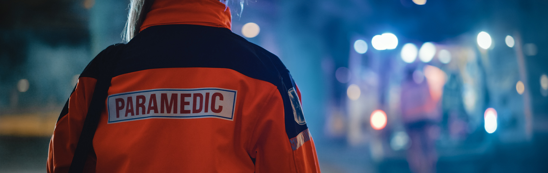 image of back of paramedic working on scene wearing a bright orange reflective jacket that says paramedic on the back. The head and lower part of the body are not visible and the background is blurred out