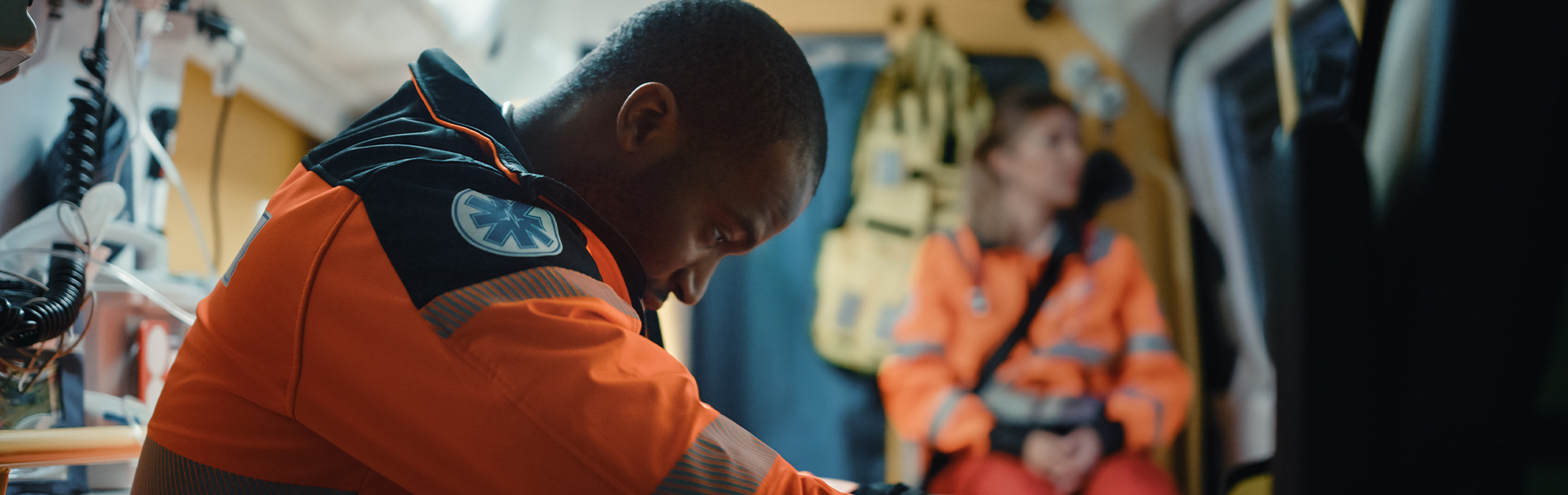 black male paramedic wearing high-visibility orange. He's hanging his head low while sitting in an ambulance