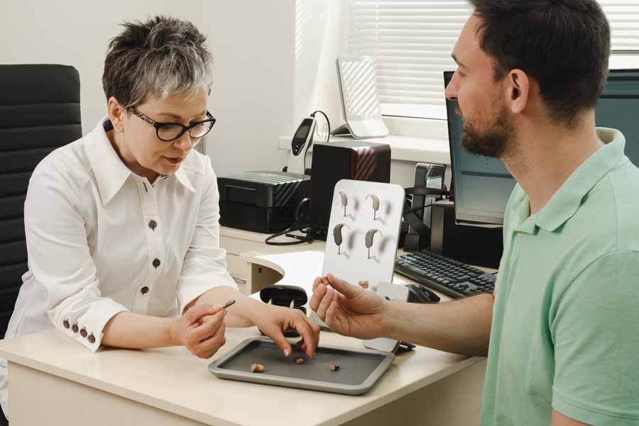 Man trying modern compact hearing aids at clinic. Audiologist consulting at hearing rehabilitation clinic to select digital device for middle age man. Doctor consulting patient