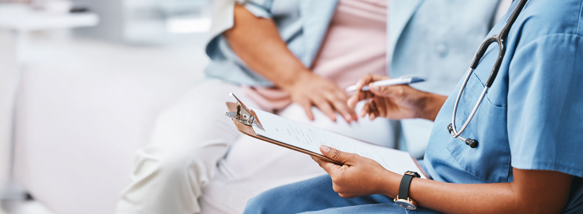 A healthcare provider in scrubs writes notes on a clipboard during a patient visit.