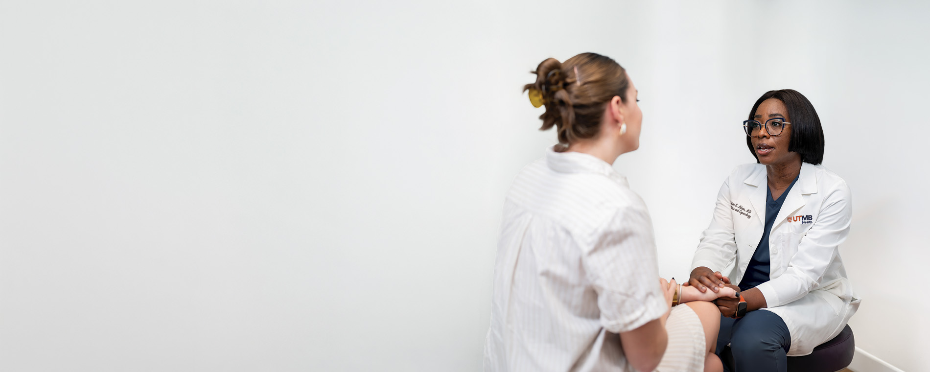 Clinician in a white coat seated across from a patient in an exam room, holding the patient’s hands during a conversation.