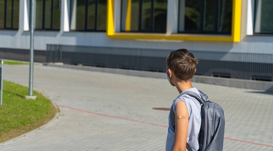 School‑aged child wearing a backpack walks toward a school building, with a small adhesive bandage visible on the upper arm, suggesting a recent vaccination before returning to class