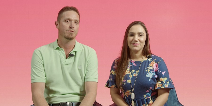 Two adults sit side by side on stools against a pink gradient background, facing forward. One wears a light green polo shirt, and the other wears a dark blue floral dress, suggesting an interview or filmed conversation setting.