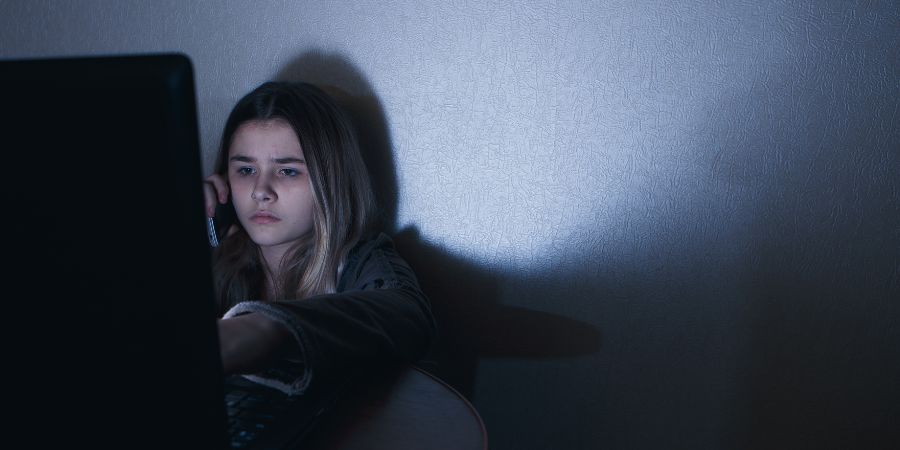 Child sitting alone in a dimly lit room using a laptop and holding a phone, with the screen glow illuminating the surrounding wall, illustrating online activity in a private space