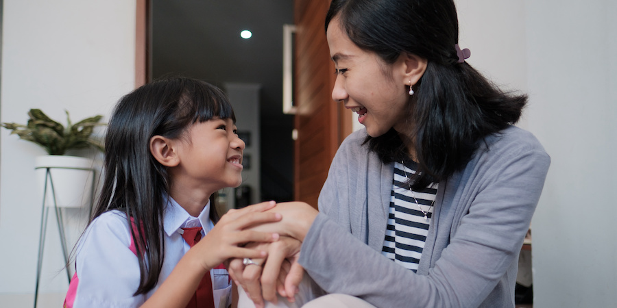 Asian daughter wearing a school uniform and backpack sitting to the left of her mother. She is holding her mother's hand and they are conversing and smiling looking at each other