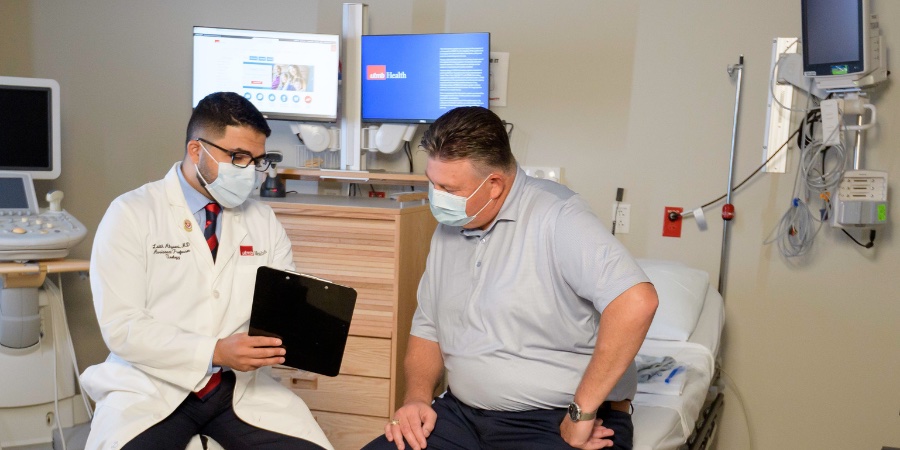 Health care provider wearing a white lab coat and face mask reviews information on a tablet with a seated patient in an exam room, with medical equipment and a hospital bed visible nearby