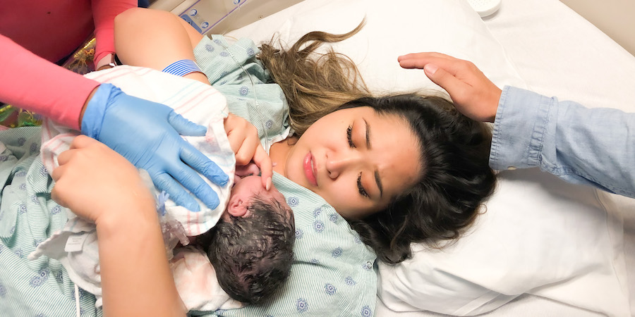 A newborn lies on the chest of a parent on a hospital bed, while gloved medical staff support the baby during moments immediately following childbirth