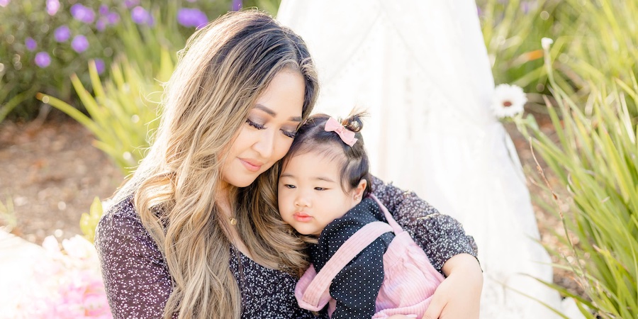 An adult sits outdoors holding a young child in their arms. Both wear casual clothing. The background includes greenery, soft sunlight and light-colored fabric, suggesting an outdoor portrait setting.