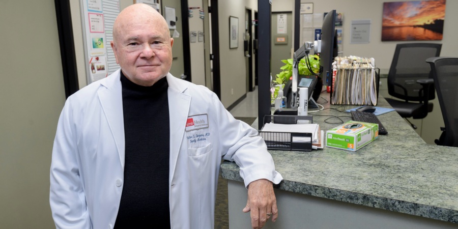 Dr. Victor Sierpina stands at a clinic reception desk at UTMB wearing a white lab coat, with a patient care area visible in the background