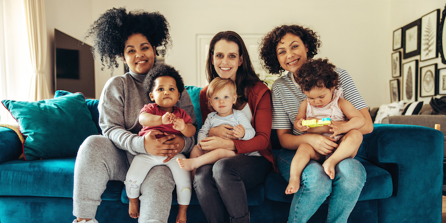 three moms sitting on a jewel tone turquoise-colored couch holding their children who all appear to be 18 months old or younger