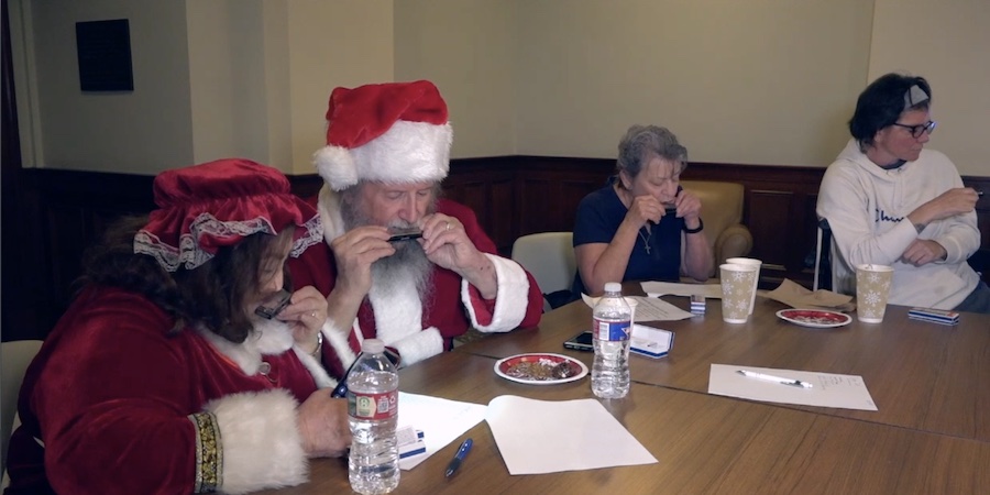 Several people seated around a table play harmonicas during a support group meeting, with papers, water bottles and cups on the table. Two participants wear Santa and Mrs. Claus holiday attire