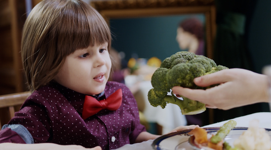 a young child wearing a button down shirt and a bow tie seated at a table, gazing at a head of broccoli being presented to them.