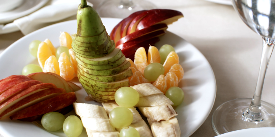 white plate topped with sliced apples, bananas, peeled oranges and full grapes with crystal glass situated to the right of it