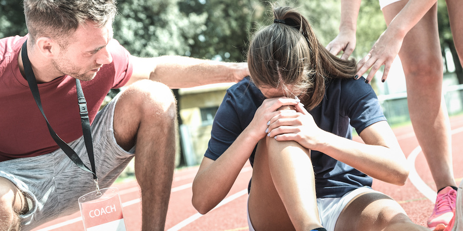 A student-athlete sits on a running track holding a knee while two adults kneel nearby and provide assistance. The setting appears to be an outdoor track during a sports practice or event.