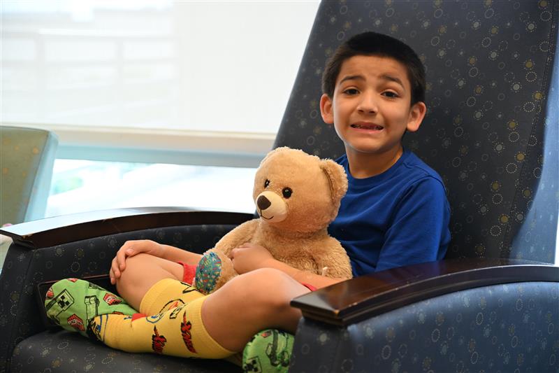 Young child seated in a padded clinic chair, holding a teddy bear, wearing a blue shirt and colorful socks, inside a medical room with soft lighting and patterned furniture