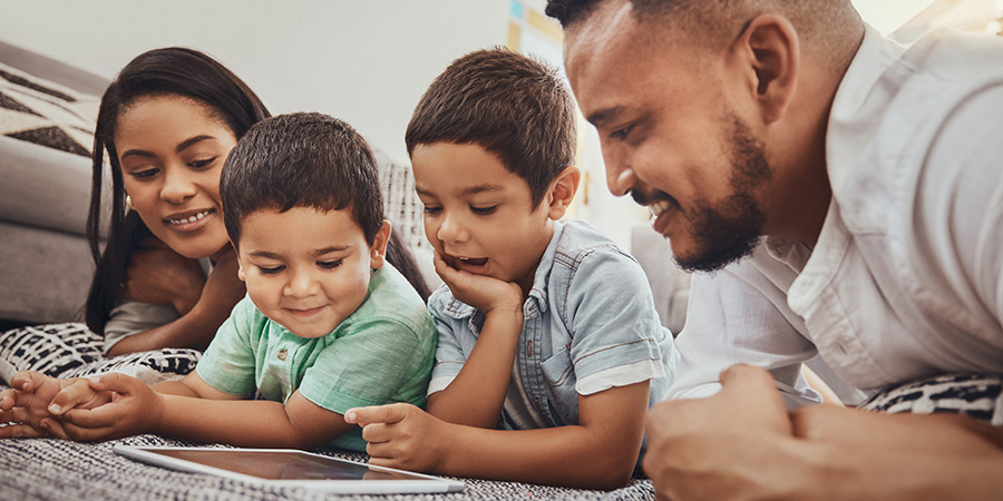Two adults and two children lie on the floor together while looking at a tablet device, with the screen positioned in front of the group in a home setting