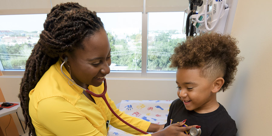black female care provider wearing a yellow shirt is using a stethoscope on a young, male pediatric patient wearing a black shirt. She is listening to his heart in a clinic room with open windows behind them. Trees are visible through the windows