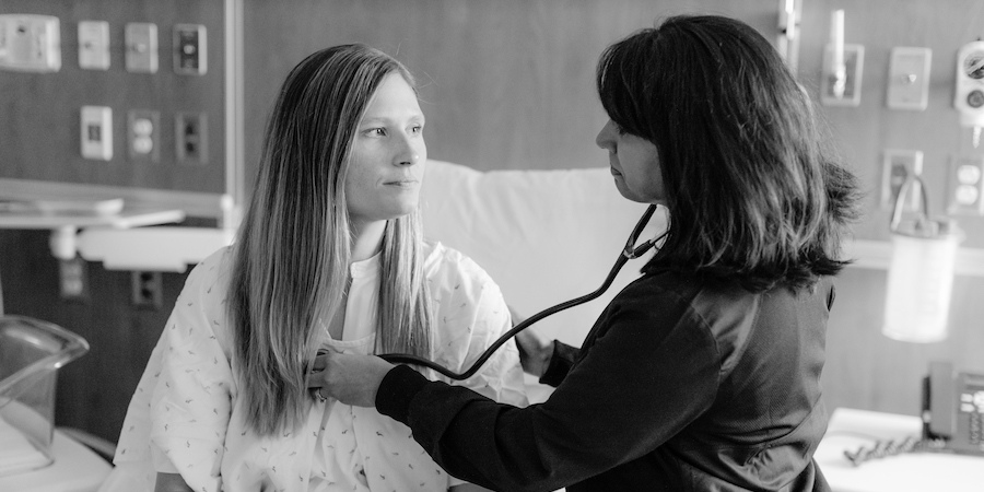 Health care provider uses a stethoscope to listen to a seated patient’s chest during a hospital visit, with medical equipment and monitors visible in the background, illustrating cardiovascular care during pregnancy or postpartum care