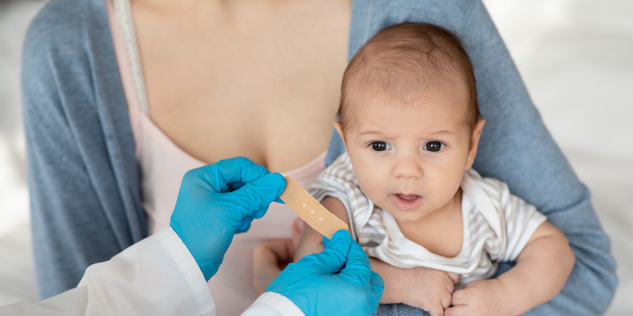 two hands in blue gloves placing a bandaid on a baby's arm. Baby is weaering a white and taupe striped onesie and is being held by a woman wearing a blue cardigan and a pink camisole