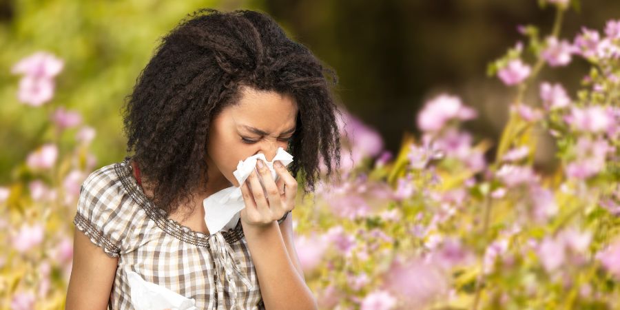 A person stands outdoors among blooming flowers while holding a tissue to their nose, with greenery and pink blossoms visible in the background