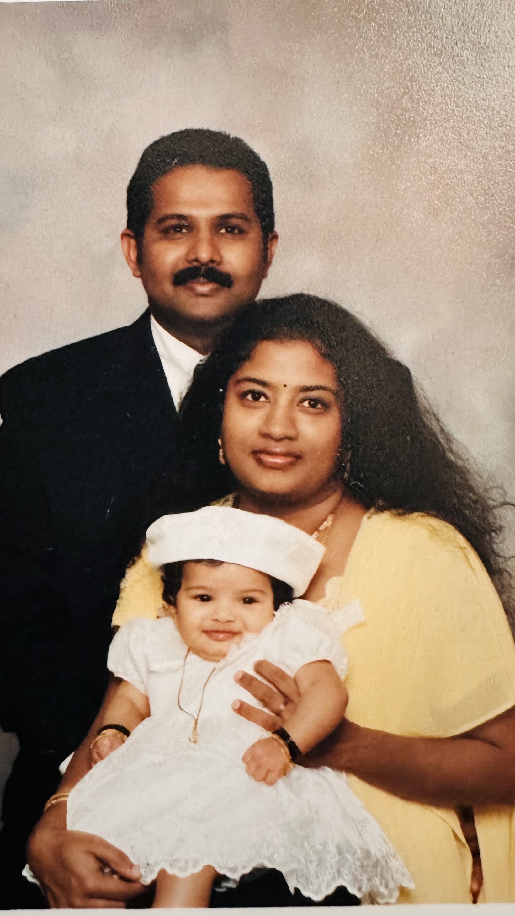 image of father, mother and infant. the male is standing in the back wearing dark clothes and smling, mother is wearing yellow clothing and holding baby girl in white dress