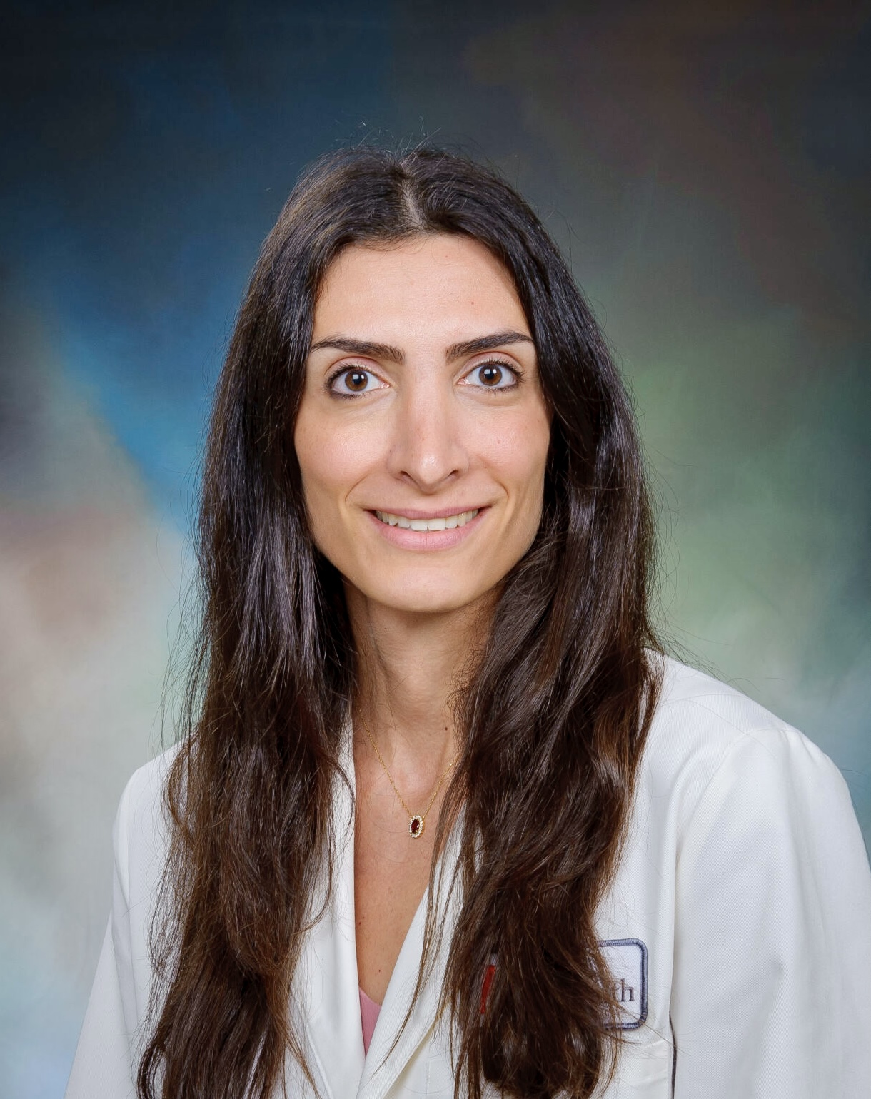 headshot of female physician with long brown hair split down the middle, wearing a white coat and smiling 