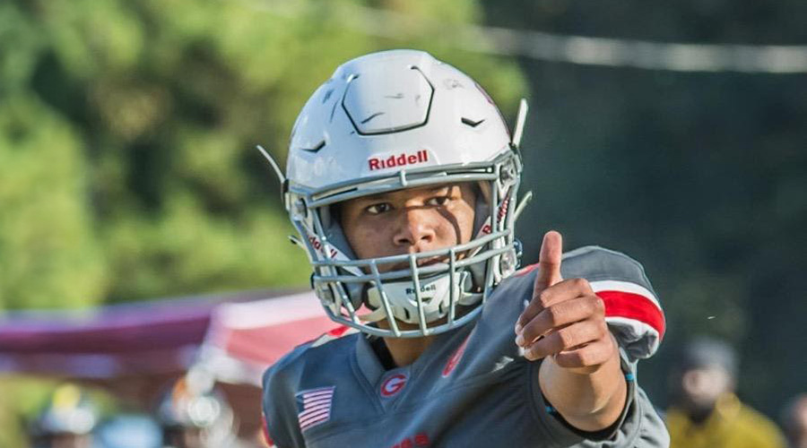 A football player wearing a helmet and uniform stands on a football field and raises a thumb, with blurred spectators and field elements visible in the background