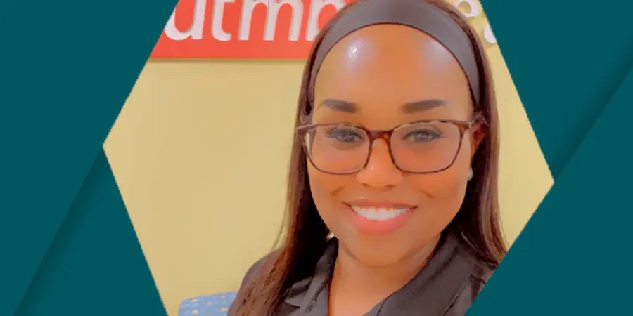 Portrait of a health care employee wearing glasses and a dark top, smiling at the camera inside a UTMB Health clinic, shown against a teal graphic background