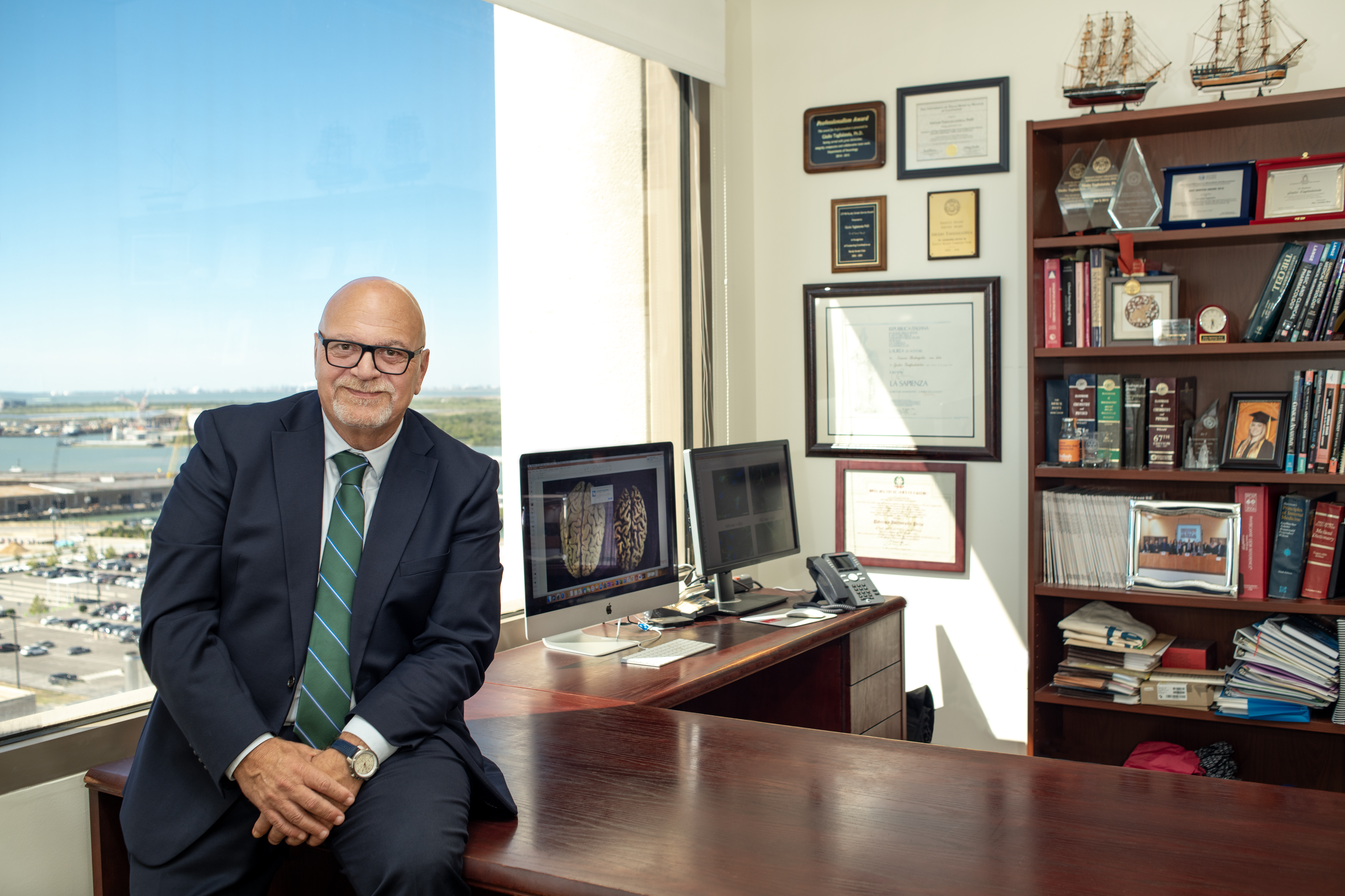 cacuasian man wearing glasses and a suit with a green striped tie leans on his desk in front of monitors that images of the brain. his degrees and book case is in the background