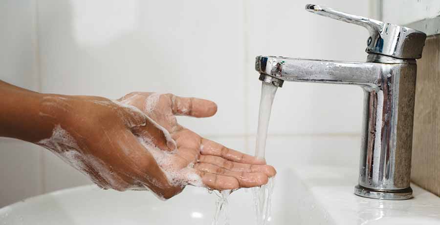 Close-up of hands covered in soap lather being rinsed under running water from a sink faucet, illustrating proper handwashing over a white basin