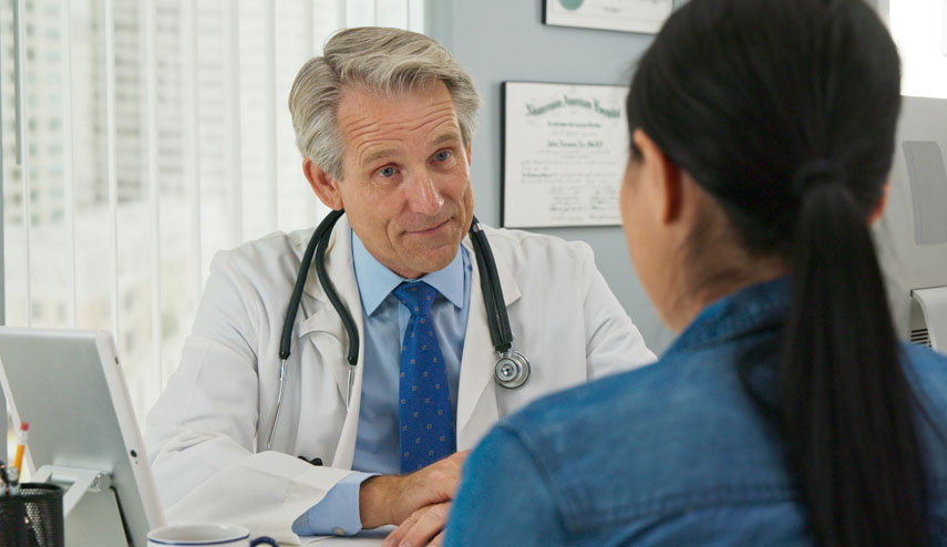 Male doctor seated behind table, speaking with female patient about her medical history