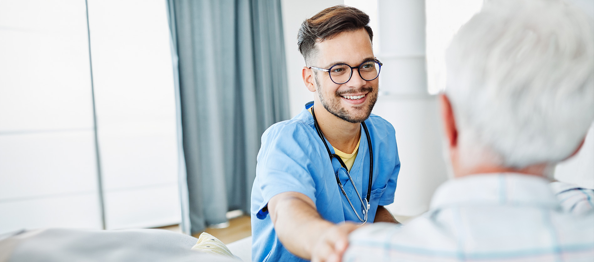 A smiling healthcare provider in scrubs speaks with an older patient during a consultation.