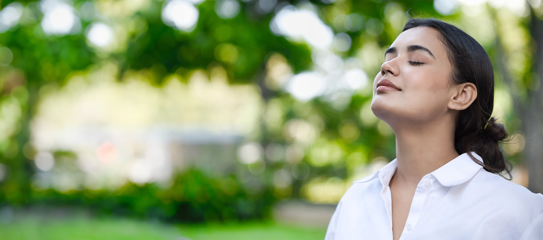 A woman in a white shirt stands outside, eyes closed, breathing deeply. She looks peaceful, surrounded by green trees in the background.