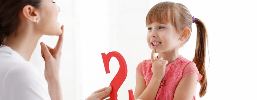 A young girl smiles while pointing to her mouth during a speech therapy session with a therapist holding the letter S.