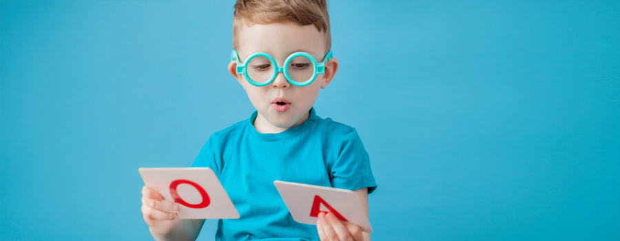 A young boy with glasses practices vowel sounds while holding letter flashcards during speech therapy.