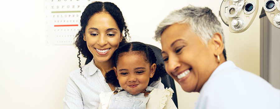 A smiling parent and child meet with an eye doctor during a pediatric eye care visit, reviewing results together.