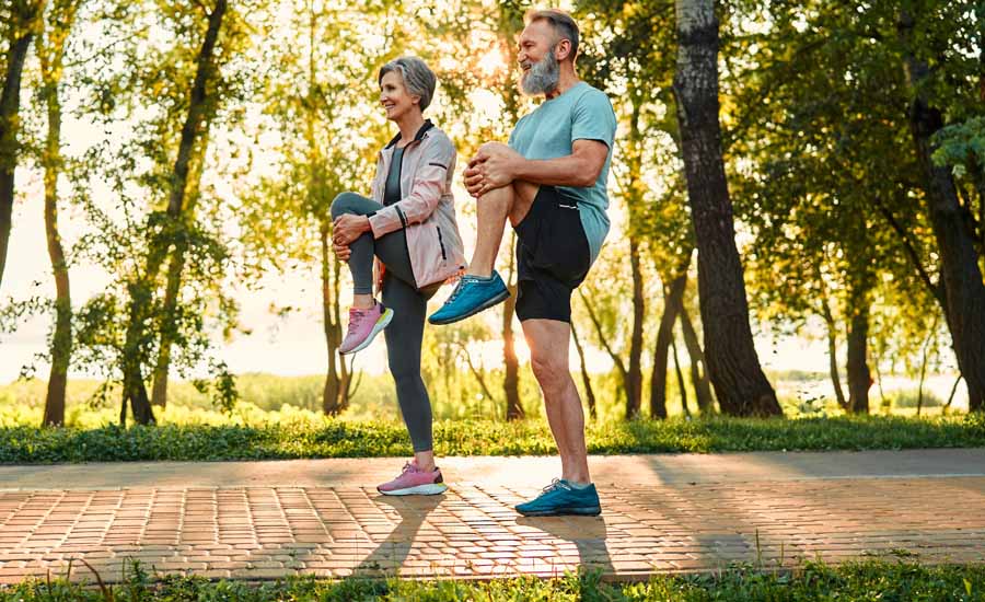 Side view of two healthy seniors standing on pathway in park and doing stretching exercises for legs
