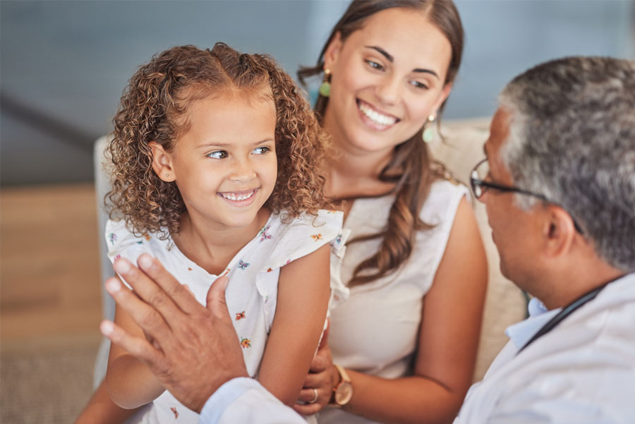 A young girl smiles while a doctor speaks with her and her mother during a pediatric nephrology exam.
