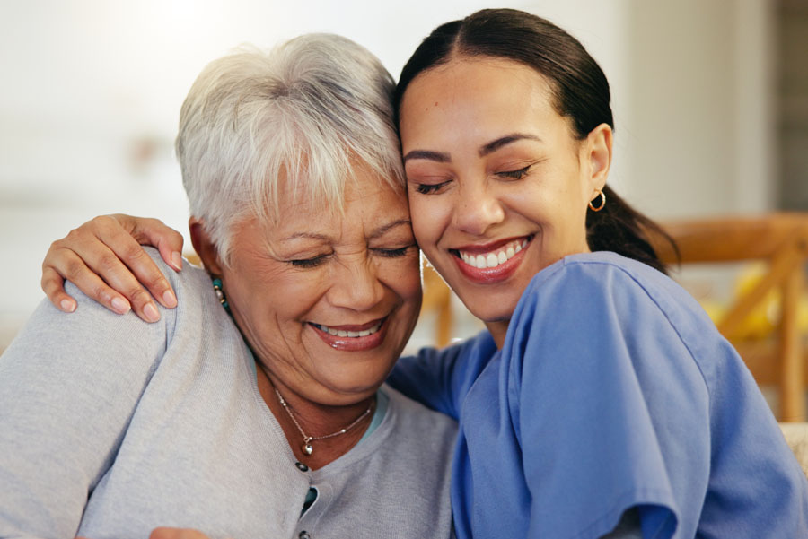 A smiling nurse and older adult patient share a warm hug while sitting on a couch.