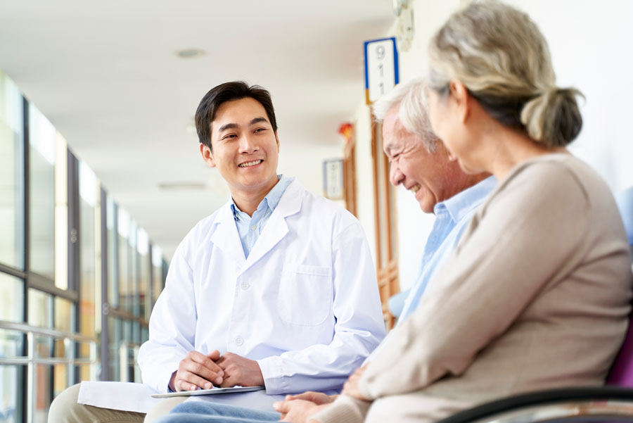 A doctor smiles while speaking with two older adult patients seated in a medical facility hallway.