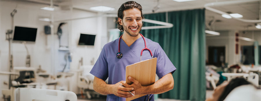 A smiling nurse in scrubs holds a clipboard while speaking with a dialysis patient in a treatment center.