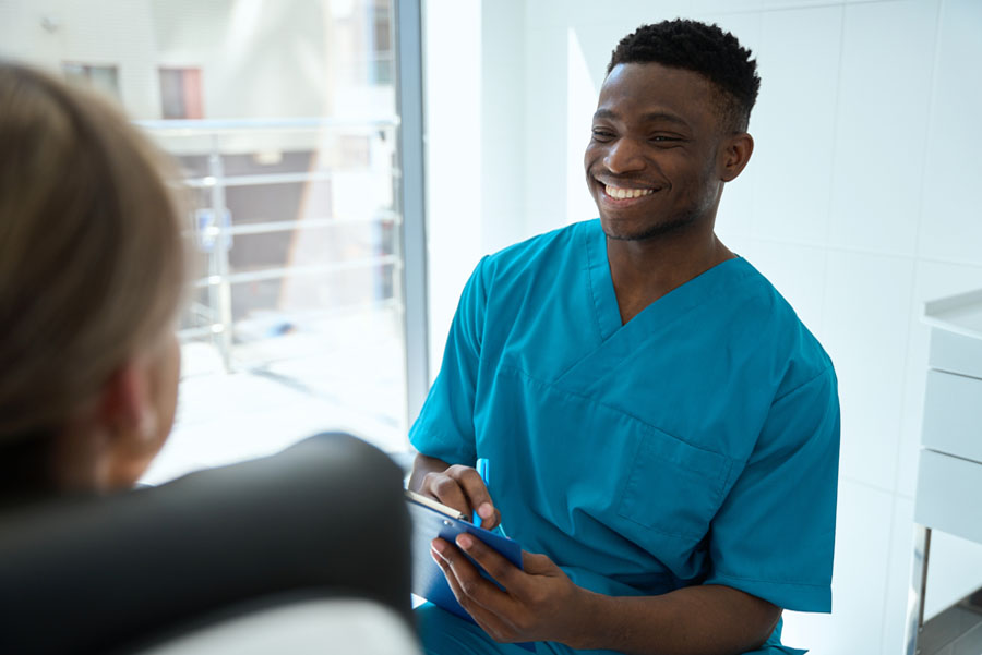 A smiling nurse in blue scrubs holds a tablet while speaking with a patient in a clinic room.