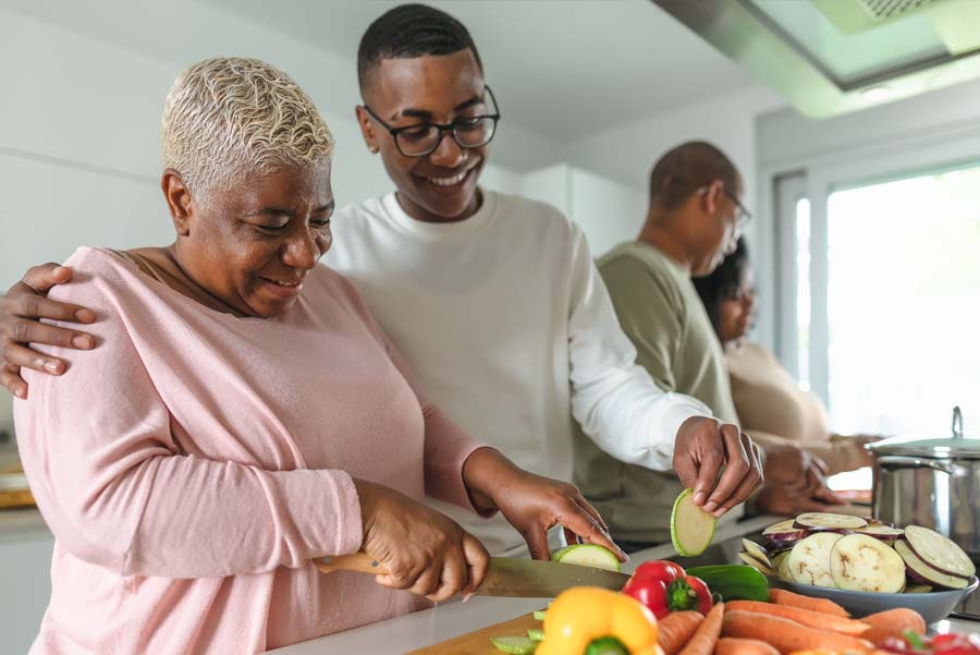 A woman smiles while chopping vegetables in a kitchen with her family, preparing a healthy meal together.