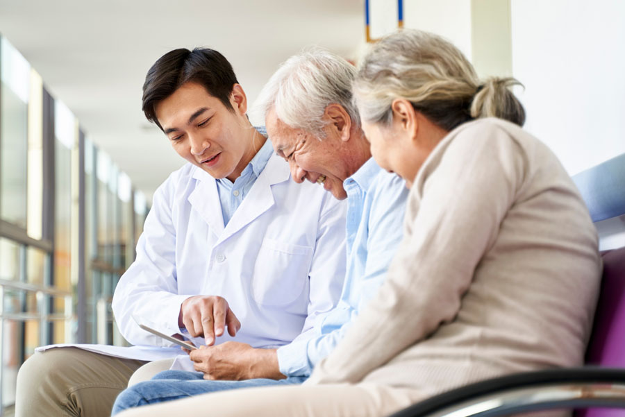 A doctor shares information on a tablet with two older adult patients seated in a clinic hallway.
