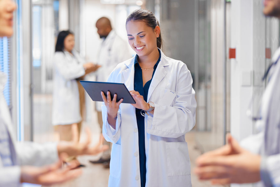 A female doctor stands in a hospital hallway smiling at a tablet while colleagues speak nearby.