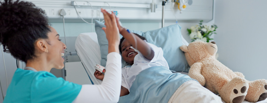 A young child smiles and gives a high-five to a nurse while lying in a hospital bed during dialysis care.