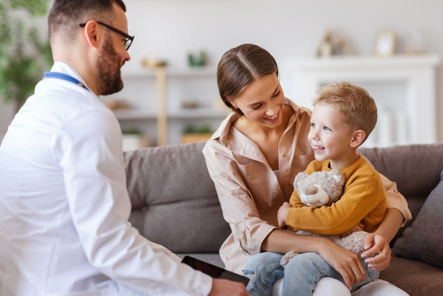 A doctor smiles while speaking with a mother and young boy, who is holding a stuffed animal on the couch.