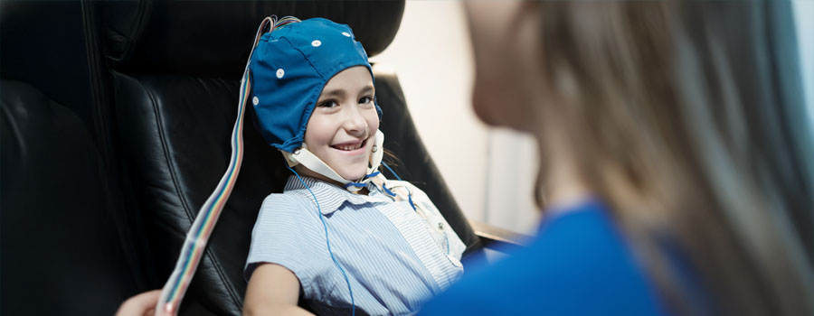 A smiling child is wearing a blue EEG cap while undergoing neurodiagnostic testing, with a healthcare provider nearby.